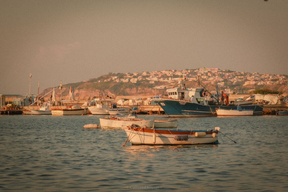 Small fishing boats and larger vessels docked in a harbour on Procida Island, with a backdrop of hillside homes and a calm, golden sea. This image perfectly captures the essence of a Procida travel guide.
