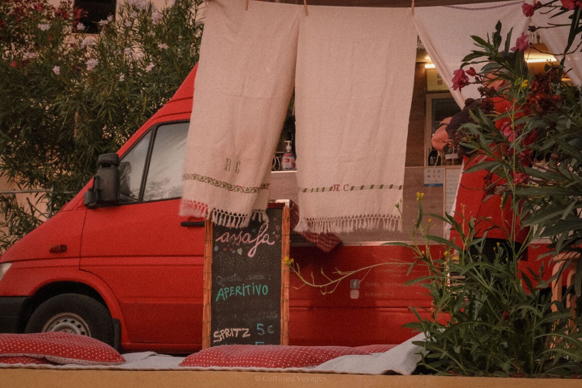 A red food truck partially hidden behind embroidered tablecloths hung to dry, with plants and a small chalkboard menu listing items like aperitifs and spritz for โฌ5, adding to Procida's rustic charm.