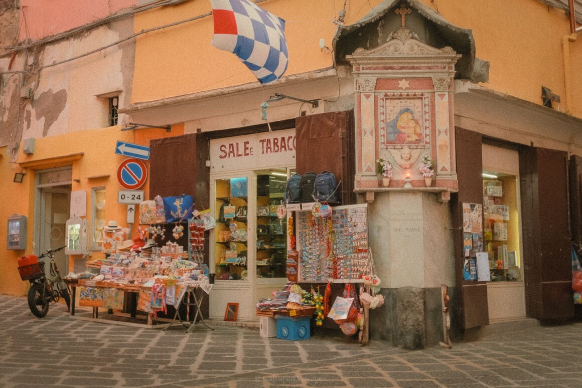 A quaint shop in Procida displaying souvenirs, trinkets, and postcards outside, alongside a small shrine decorated with flowers and a candle, offering a glimpse into local culture.