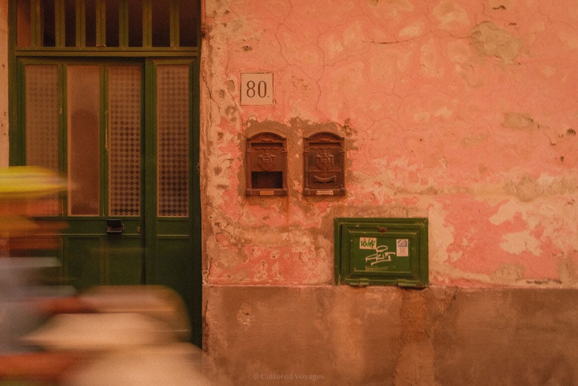 A weathered pink wall featuring two vintage brown mailboxes and a green utility box, with blurred motion in the foreground, showcasing Procidaโs authentic charm for a Procida travel guide.