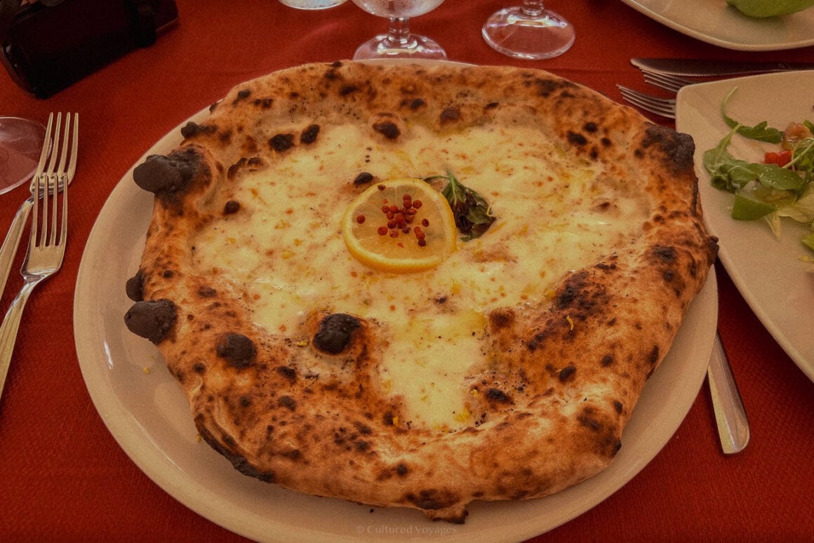 A close-up of a wood-fired pizza topped with melted cheese, a lemon slice, and pink peppercorns, served on a white plate with accompanying cutlery and side dishes on a red tablecloth.