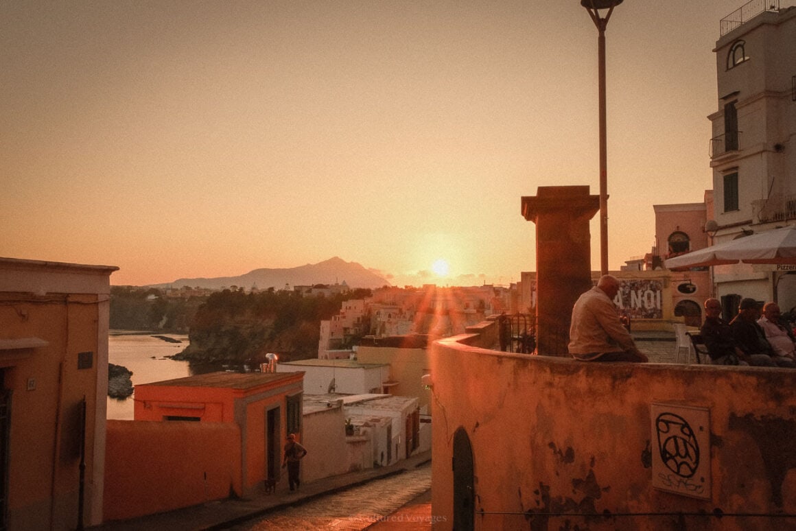 A sunset view of a picturesque coastal village on Procida Island, Italy. The sun casts a golden glow over colourful houses and a distant silhouette of mountains, while a few people sit and chat near a curved wall.