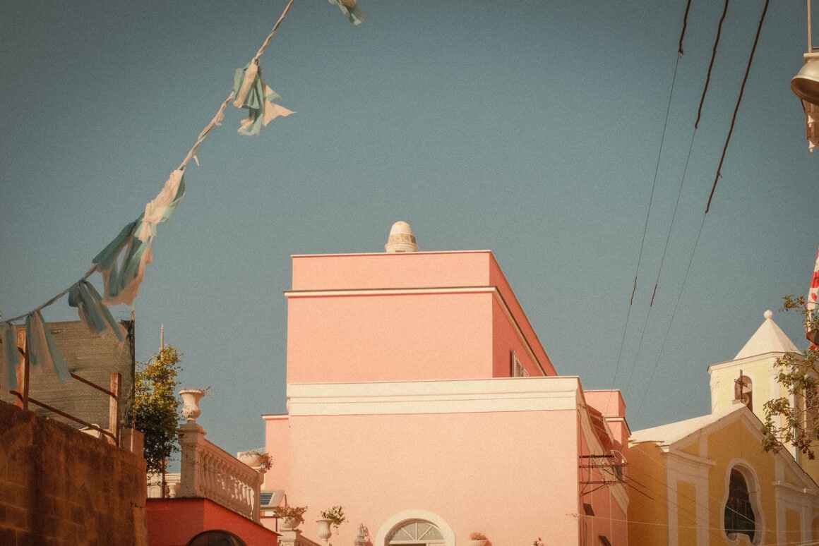 A pastel pink building with decorative white accents on Procida Island, surrounded by festive strings of flags, set against a clear blue sky.