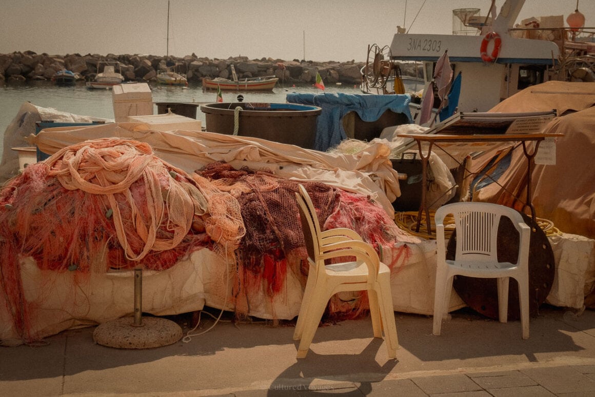 Piles of colourful fishing nets, plastic chairs, and equipment scattered along the harbourfront, with small fishing boats anchored in the background.