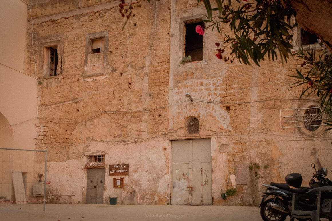 The weathered facade of an old stone building in Procida, labelled as "Museo" with faded signs and iron doors, surrounded by blooming flowers and a parked scooter.