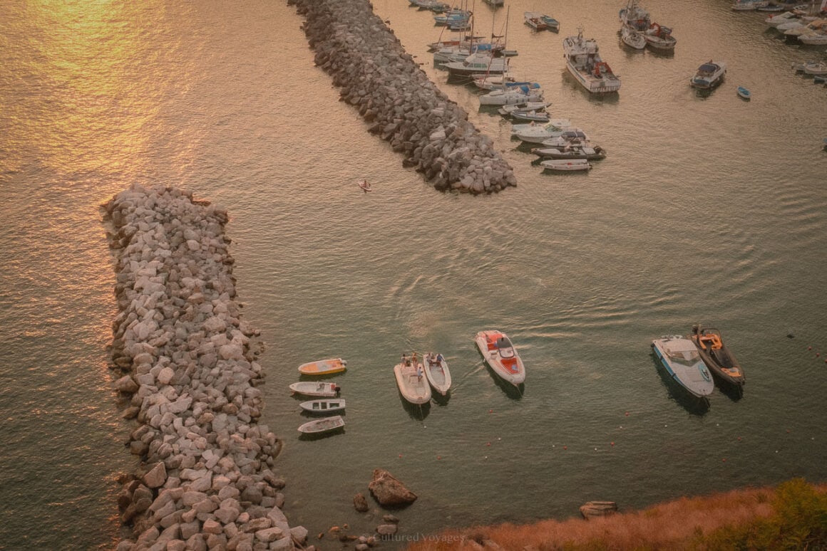A harbour view at sunset featuring small boats gently floating in the water, bordered by rocky breakwaters and shimmering in the golden light of Procidaโs coastline. This image is ideal for a Procida travel guide.