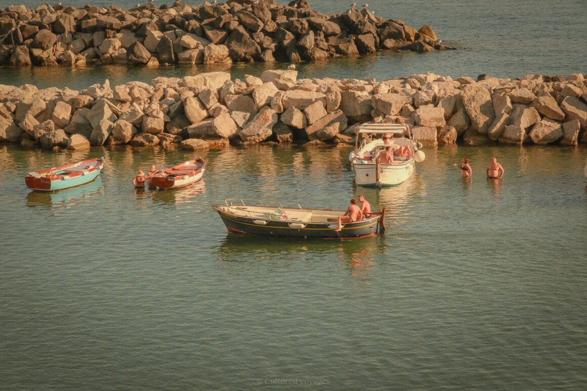 A close-up view of small fishing boats near a rocky seawall, with a few people enjoying the water under the warm glow of the afternoon sun.