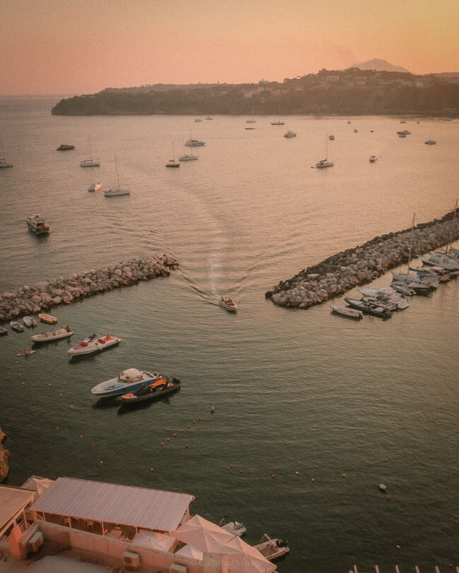 An elevated view of a harbour at sunset, with boats scattered across the calm water and a breakwater leading out to the horizon, framed by the golden glow of the evening.