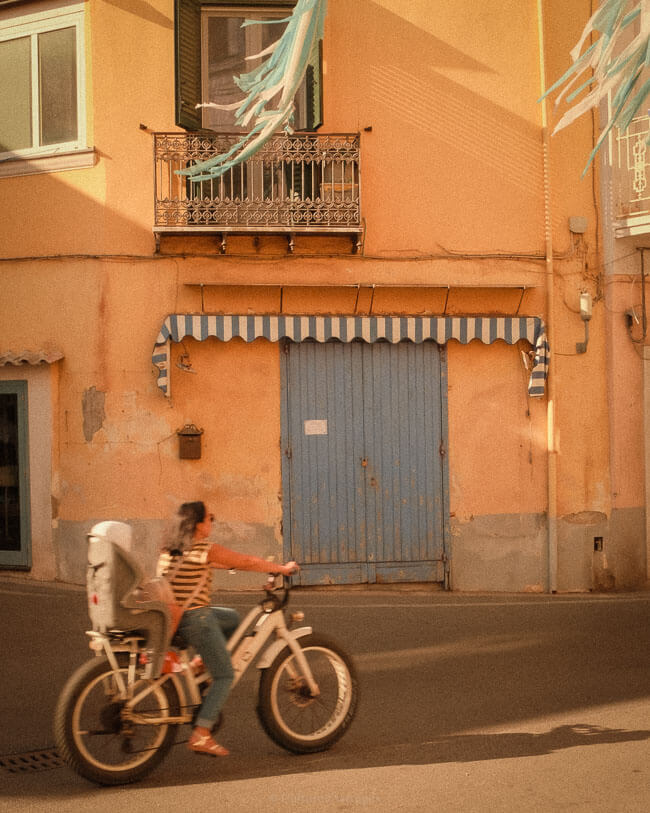 Woman cycling past a warm-toned building with blue doors, striped awning and traditional balcony in Ischia, Italy.