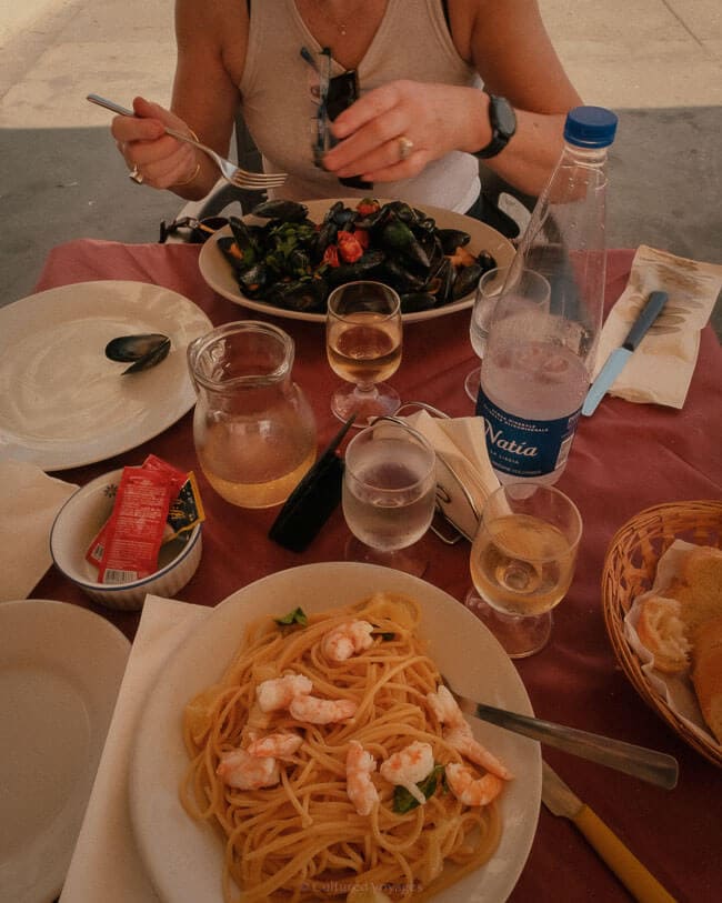 A table set for a seaside meal featuring plates of prawn pasta and fresh mussels, accompanied by glasses of white wine, water, and bread. The simple yet delicious spread highlights local cuisine for a Procida travel guide.