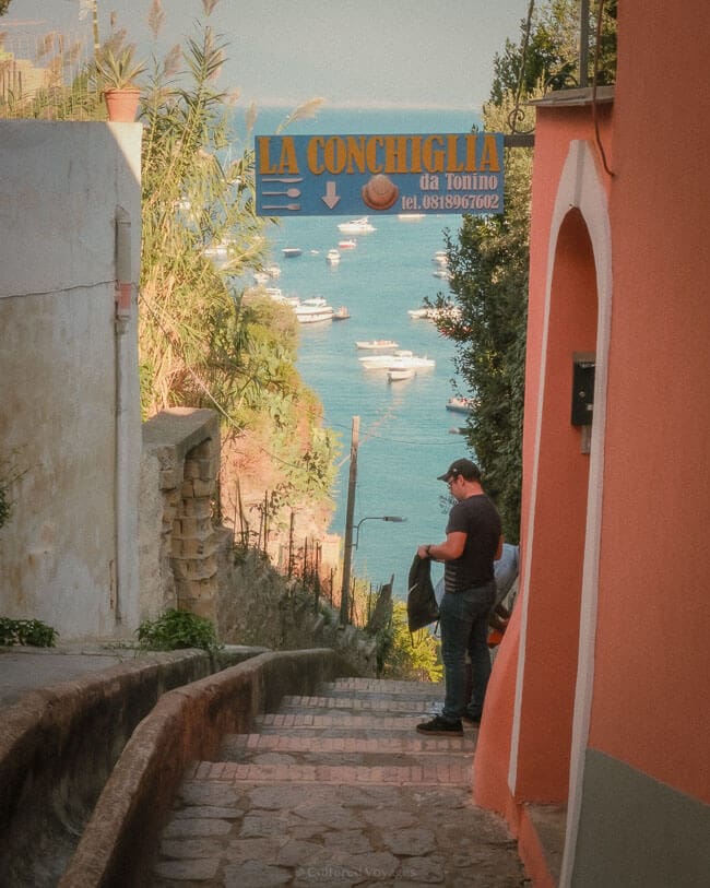 A narrow cobblestone path descending towards the sea on Procida Island, framed by colourful buildings and greenery, with a man standing near a restaurant sign reading "La Conchiglia."