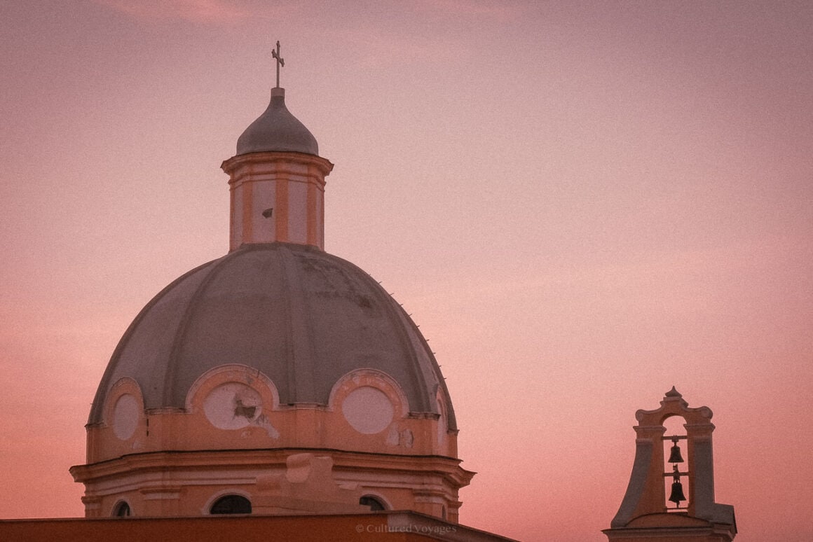 The dome of a historic church in Procida silhouetted against a pink and orange sunset sky, accompanied by a smaller bell tower with visible chimes. This image captures a peaceful moment suitable for a Procida travel guide.