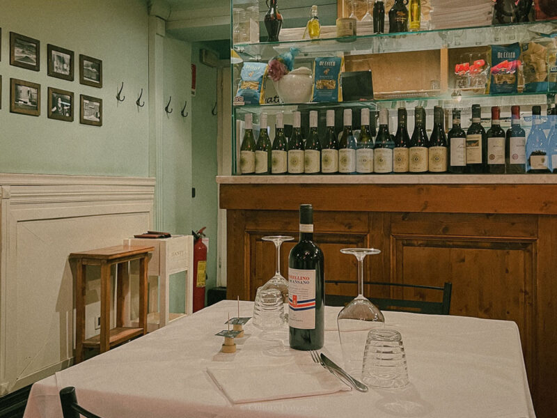 A small restaurant table set with a bottle of red wine and glasses sits in front of a shelf displaying various wine bottles. The walls are decorated with framed photos.