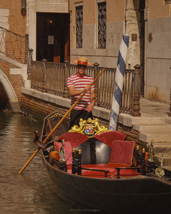 Is Venice worth visiting? A gondolier in a red-and-white striped shirt and straw hat stands beside his gondola, which features plush red velvet seating and gilded ornamentation. A blue-and-white mooring pole stands nearby, marking a docking point along the canal.