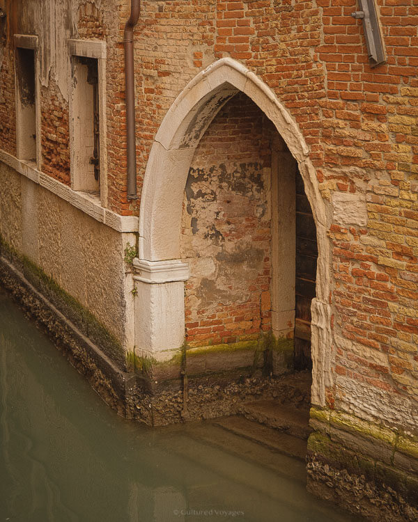 A close-up of a historic Venetian building’s arched doorway, partially submerged in canal water. The aged red brick and peeling plaster contrast with the ornate white stone arch, a reminder of the city's centuries-old architecture and its ongoing battle with rising water levels.