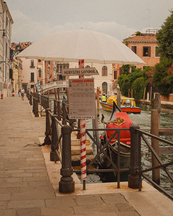 Is Venice worth visiting? A gondola service station along a Venetian canal, marked by a striped red-and-white pole and a large white umbrella. A sign reading "Servizio Gondola" provides information for tourists, while a gondola with vibrant red seating waits in the water.