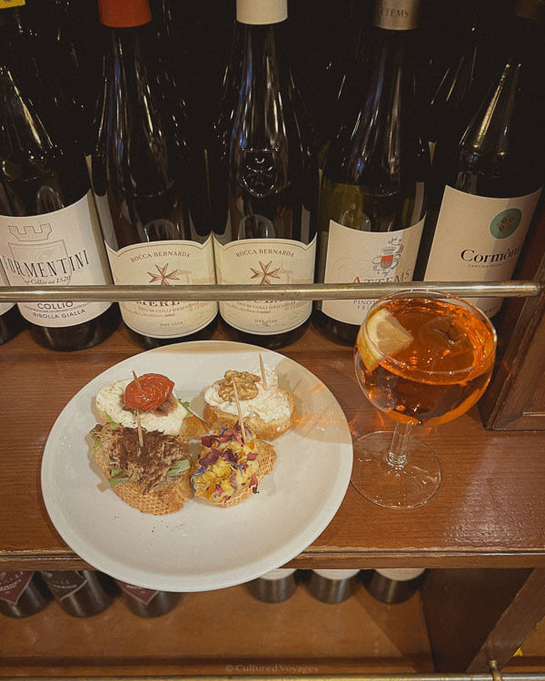A plate of Venetian cicchetti, small appetisers on toasted bread, sits on a bar counter alongside a glass of Aperol Spritz. Behind the plate, a row of fine Italian wine bottles adds to the rustic charm of the traditional Venetian bacaro setting.