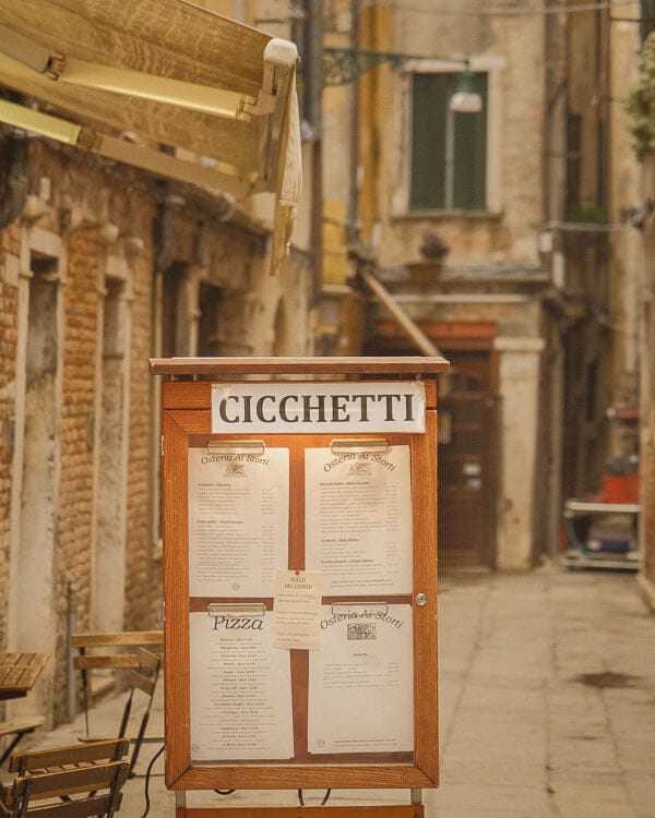 A wooden menu board displaying a selection of Venetian cicchetti and pizza stands outside a cosy trattoria. The surrounding alleyway, with its warm-toned bricks and vintage awnings, captures the essence of a hidden Venetian dining spot.
