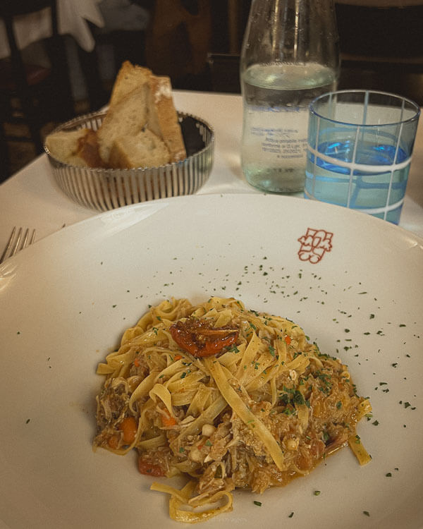 Is Venice worth visiting? A plate of fresh tagliolini pasta with crab, garnished with herbs and cherry tomatoes, is served at a Venetian restaurant. A basket of rustic bread, a bottle of water, and a glass with a blue rim complete the simple yet inviting dining setup.
