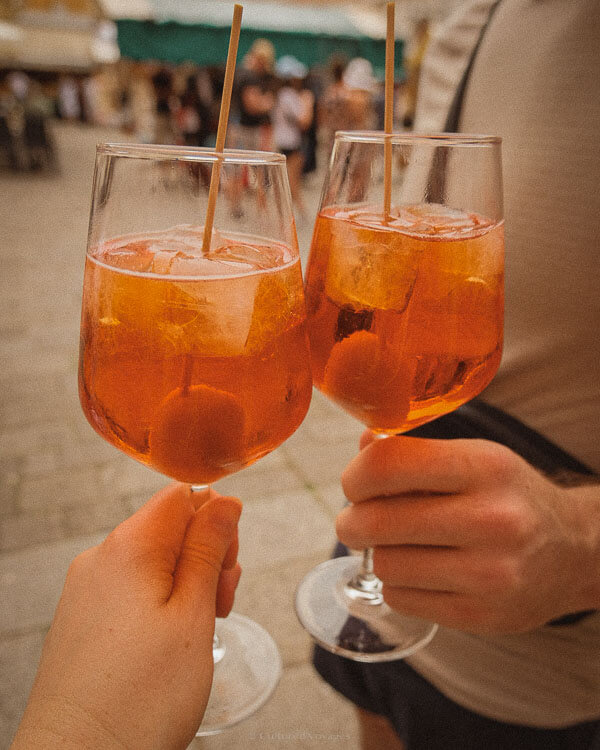 Two people toast with glasses of Aperol Spritz in a lively Venetian square. The golden-orange cocktails, garnished with olives, glisten under the soft afternoon light, capturing the essence of Italy’s aperitivo culture.