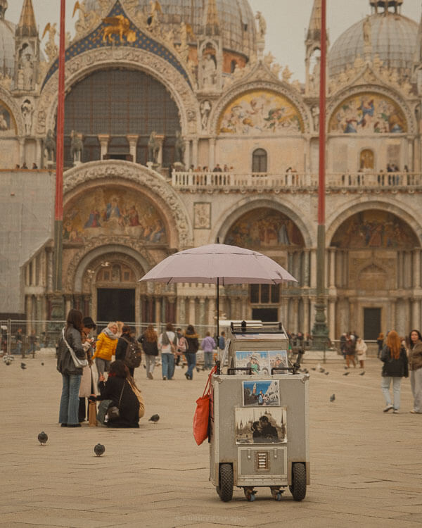 A small street vendor cart with a lavender umbrella stands in front of St Mark’s Basilica in Venice. The cart is filled with postcards and souvenirs, while tourists walk through the square, surrounded by pigeons. The basilica’s intricate mosaics and domes serve as a stunning backdrop.