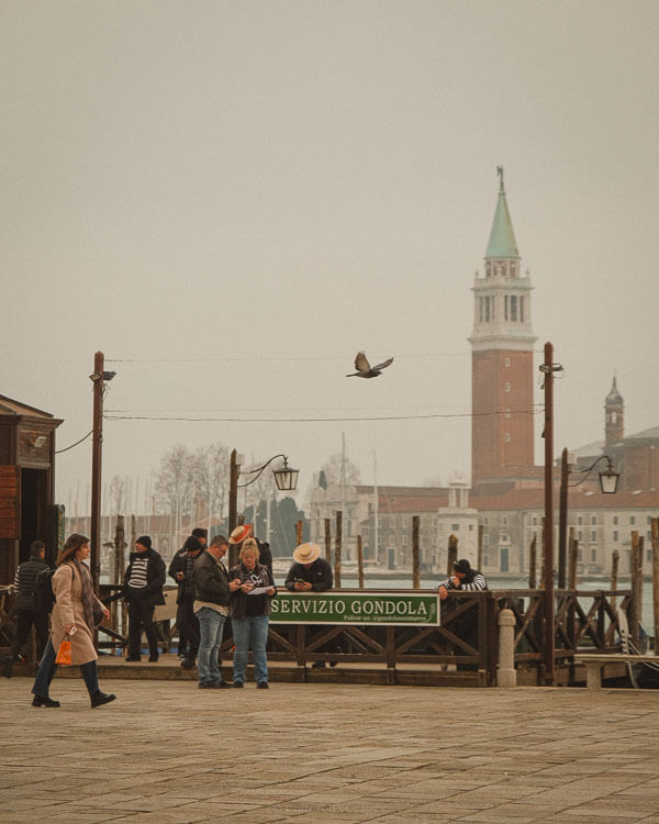 A misty morning at a Venetian gondola station, where people gather near a green "Servizio Gondola" sign. Gondoliers in striped shirts prepare for the day as a pigeon flies overhead. In the background, the bell tower of San Giorgio Maggiore rises above the lagoon, partially obscured by the fog.