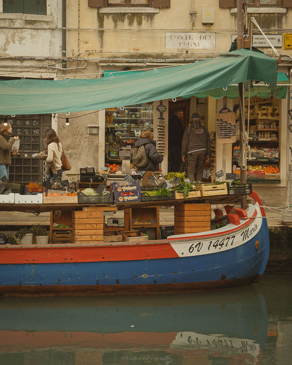 A floating fruit and vegetable market in Venice, set up on a brightly painted boat along the canal. Is Venice worth visiting? Fresh produce, including leafy greens and oranges, is neatly arranged in crates, while locals browse under a green canopy. A traditional Venetian grocery shop stands in the background, its entrance lined with wooden shelves filled with goods.