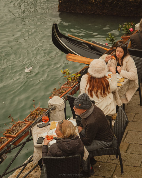 Diners enjoy a meal at a waterside café in Venice, sipping Aperol Spritz while overlooking a canal. A black gondola with polished wooden oars floats nearby, and a seagull drifts on the water. The diners are wrapped in warm coats and hats, embracing the cosy atmosphere of a winter day by the canal.