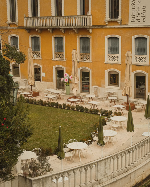 An elegant outdoor courtyard in Venice, featuring neatly arranged white metal tables and chairs surrounded by a manicured lawn. Tall, ochre-coloured buildings with arched windows and ornate balconies frame the serene space, while large folded umbrellas hint at a quiet moment between bustling café hours.