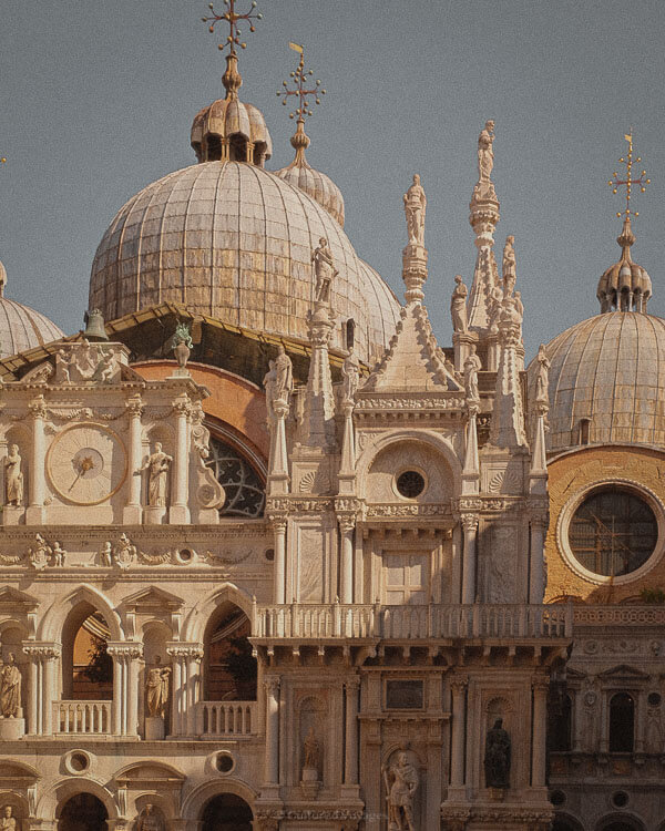 A close-up of the domes and spires of St Mark’s Basilica in Venice, showcasing its elaborate Gothic and Byzantine architectural details. Statues and ornate carvings adorn the white marble façade, with golden crosses gleaming atop the domes under the afternoon sun.