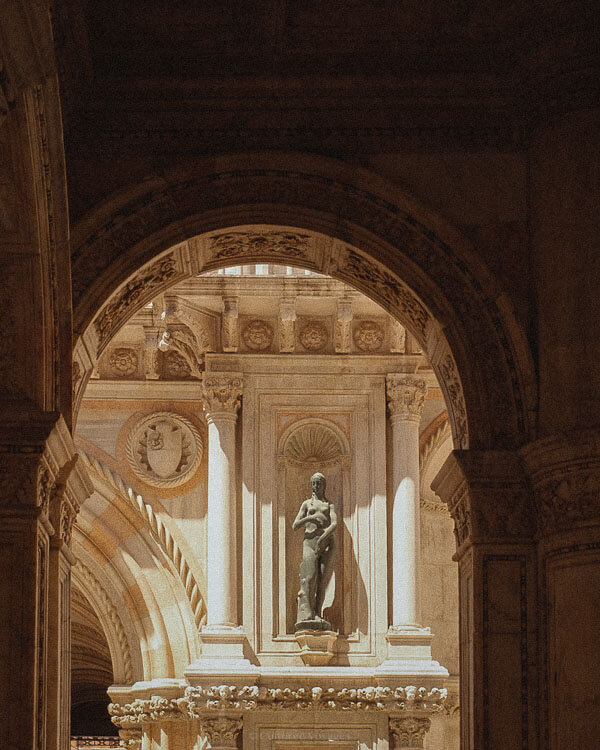 A grand arched doorway inside the Doge’s Palace in Venice frames a classical bronze statue. Sunlight filters through, highlighting the intricate stone carvings, coffered ceilings, and historical elegance of the Venetian landmark.