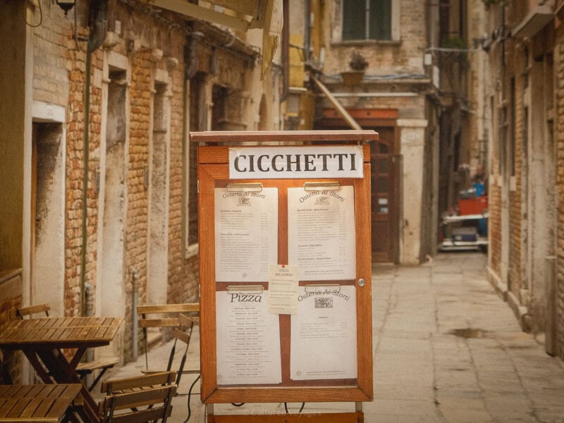 A wooden menu board displaying the word "Cicchetti" stands in a narrow Venetian alleyway, surrounded by rustic brick buildings. Small wooden tables and chairs sit to the side, hinting at a cosy dining spot.