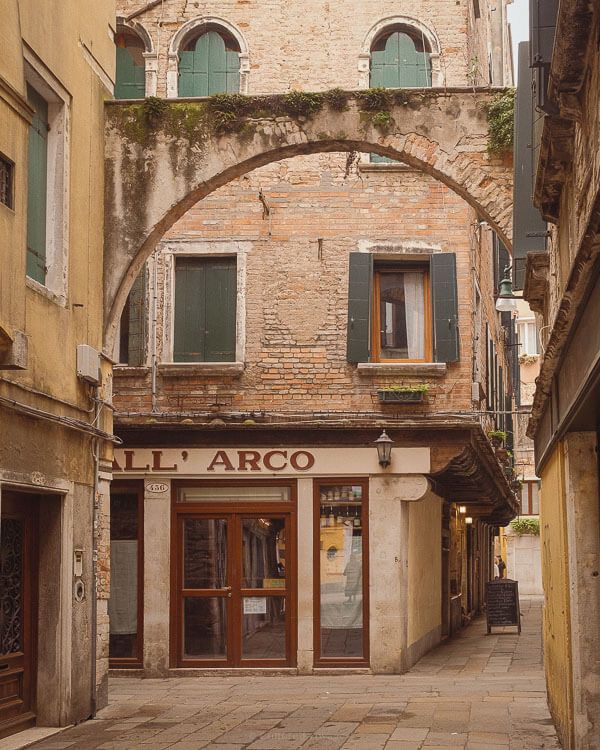 A charming bacaro named All' Arco tucked away in a narrow Venetian alley, framed by an old brick archway. The warm glow from inside suggests a welcoming place for locals and visitors to enjoy authentic cicchetti.