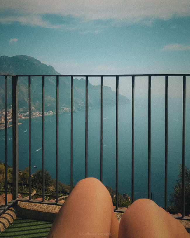 A relaxed view of the Amalfi Coast from behind a metal railing, with a pair of tanned knees in the foreground and the vast, blue sea and mountains stretching into the distance.