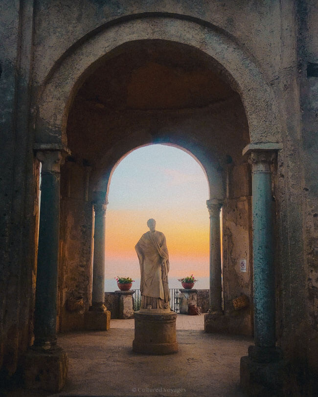 A classical marble statue framed by ancient archways at Villa Cimbrone gazes out to a glowing orange-pink sunset sky over the sea, perfectly capturing why many ask, is Ravello worth visiting.