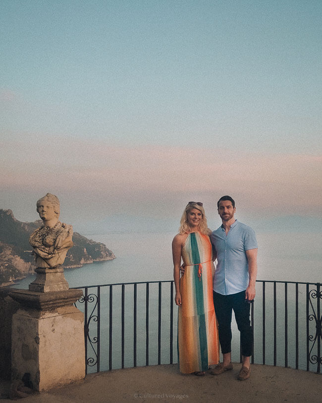 A smiling couple stands at a scenic viewpoint in Ravello during sunset, with a marble bust statue and sweeping sea views behind them.