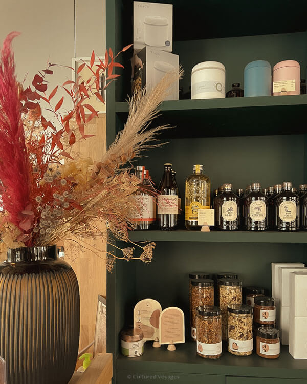 A dark green shelf in a boutique-style shop displaying an assortment of artisanal goods including bottles of oils and vinegars, jars of herbs and teas, and natural wellness products, with a large dried floral arrangement on the side.