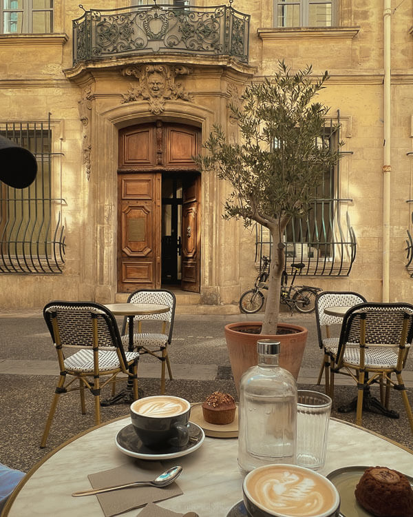 Outdoor café table in Avignon with two cappuccinos, a chocolate muffin, and sparkling water, set against a charming façade with a large wooden doorway, wrought-iron balcony, and parked bicycles.