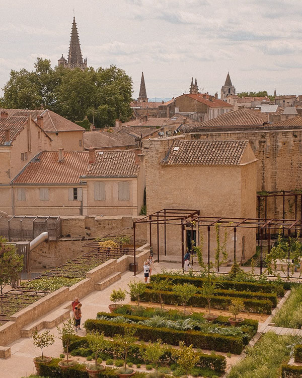 Elevated view of a neatly arranged medieval-style garden in Avignon, framed by ochre-roofed houses and distant church spires under a partly cloudy sky.