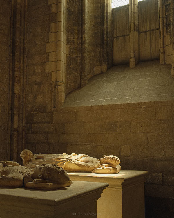 A soft-lit chapel room within the Palais des Papes, where intricately carved medieval tombs lie in the warm glow beneath a stone window.