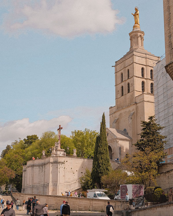 Tourists stroll past the Avignon Cathedral, its tower crowned by a golden Virgin Mary statue, beside lush green trees and medieval stone structures.