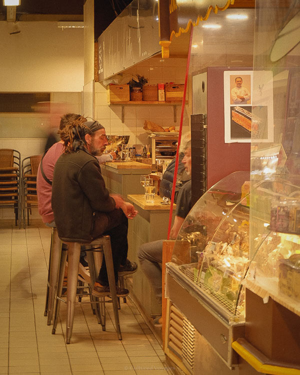 Cosy indoor bistro in Avignon where patrons sit at the bar sipping drinks, with warm lighting and shelves stacked with regional cheeses and charcuterie — a typical scene from one day in Avignon.