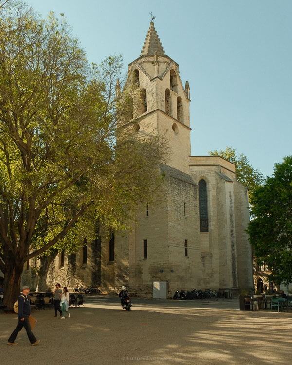 A view of a stone church tower surrounded by leafy trees, with pedestrians walking through the open square beneath.
