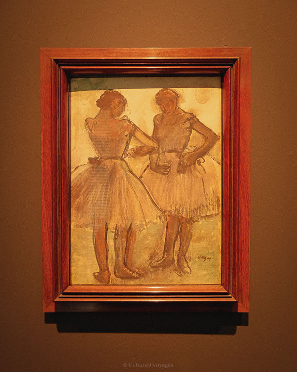 A framed pastel artwork of two ballerinas in tutus mid-conversation, displayed against a warm brown gallery wall during one day in Avignon.