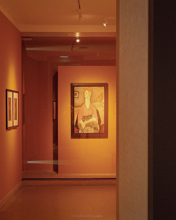 A softly lit museum corridor leading to a framed portrait of a woman with red hair wearing a patterned skirt, hanging on a terracotta-coloured wall.