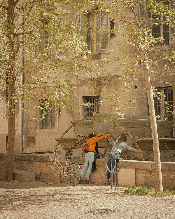 Two women leaning over a large wooden water wheel in a shaded square, surrounded by stone buildings and leafy trees.