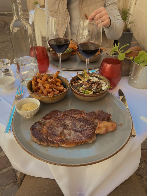 A table set for two outdoors, featuring a rib-eye steak, sweet potato fries, salad, and two glasses of red wine, served on neutral-toned plates.