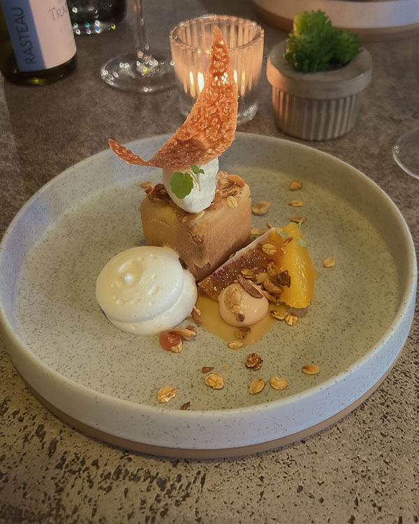 A beautifully plated dessert in a fine dining setting, featuring a sponge cake topped with whipped cream and caramel shard, surrounded by citrus, nuts and a quenelle of white mousse.