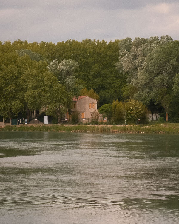 A tranquil riverside scene at sunset showing a quaint stone house nestled among dense trees along the Rhône River, reflecting peaceful countryside charm.