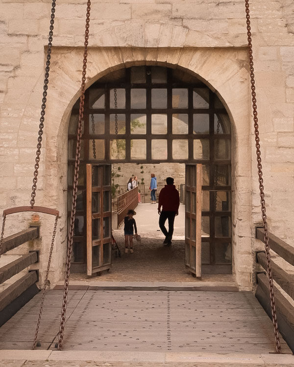 Silhouettes of visitors walking through a medieval portcullis gate and onto a bridge, framed by heavy wooden doors and thick stone walls in Avignon.